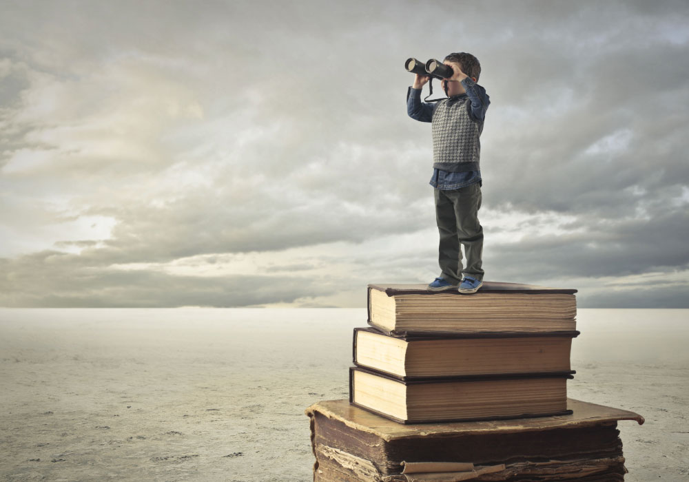 Child standing on a stack of books holding binoculars and looking into the distance under a cloudy sky.