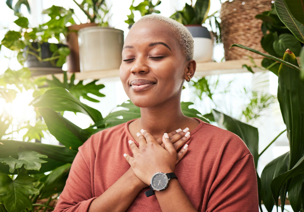 Person with eyes closed and hands on their chest, surrounded by houseplants in soft daylight.