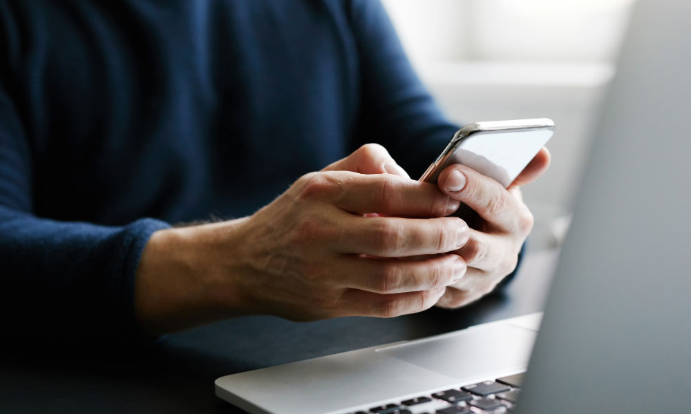 Person holding a smartphone while sitting beside a laptop, representing the elements used in everyday technology.