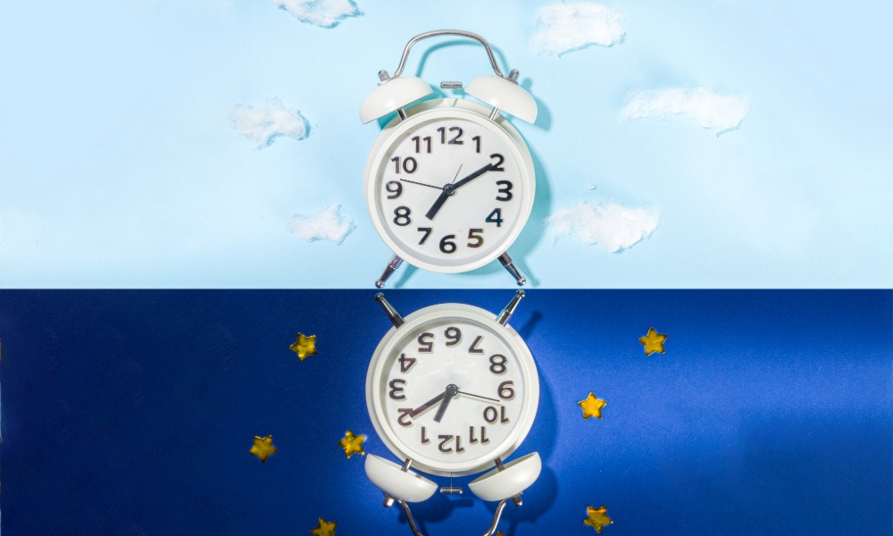 Two alarm clocks against a split background showing daytime sky above and nighttime sky below.