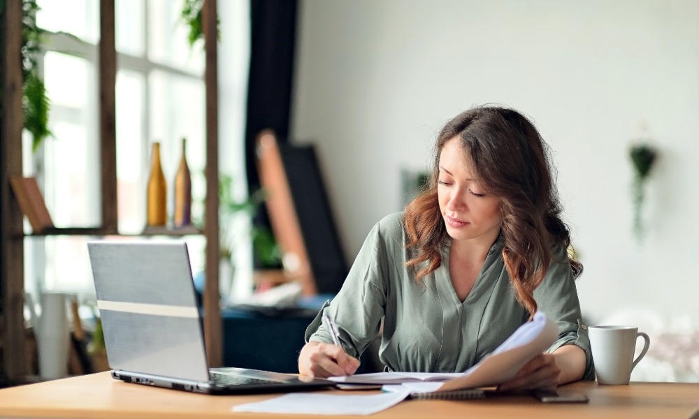 Woman sitting at a desk working with papers and a laptop in a bright home office.