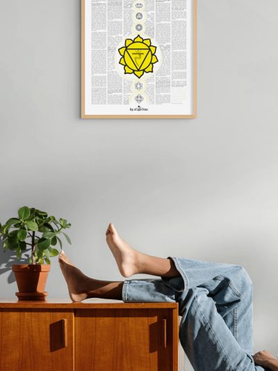 Solar plexus chakra print in a light wood frame above a wooden cabinet, with a potted plant and a reclining person resting their feet on top.